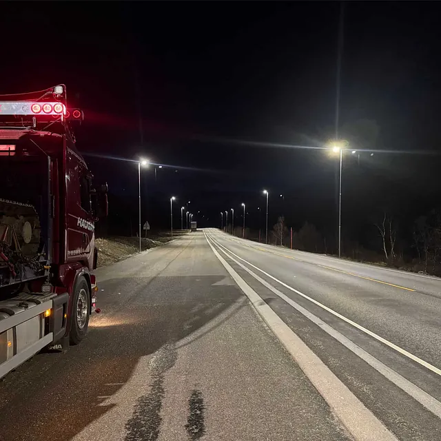 Lastebil med gravemaskin på henger står parkert på opplyst vei om natten.