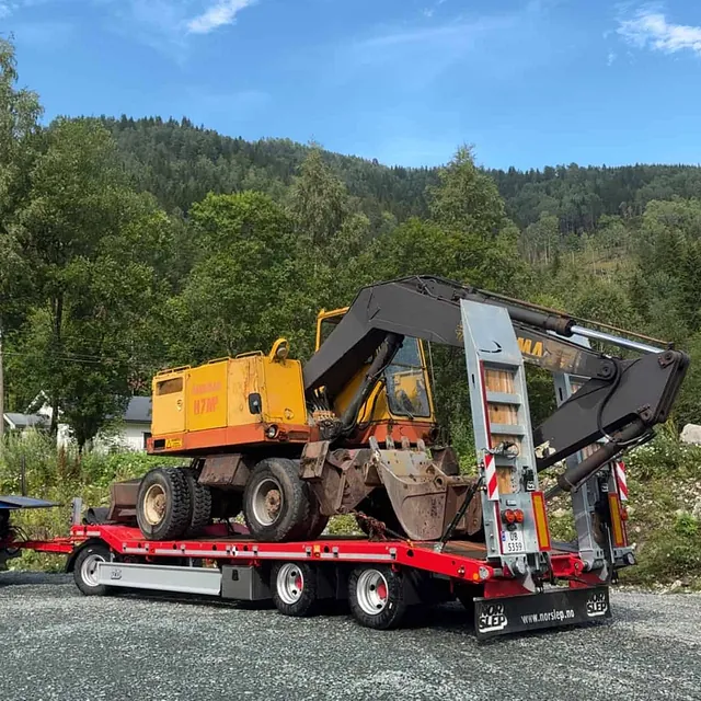 Gul og rusten hjulgraver lastet på rød maskinhenger, klar for transport på grusplass i skogkledd område.