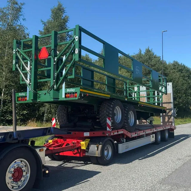 Grønn landbruksvogn fra Bailey transportert på spesialhenger, parkert på vei ved skogkant.