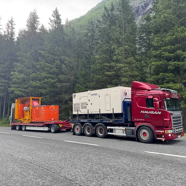 Lastebil med Skanska-utstyr og kabeltrommel parkert ved skogkledd fjellside.