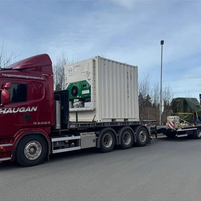 Rød Haugan-lastebil frakter stor hvit container og tildekket last på spesialhenger langs industrivei.