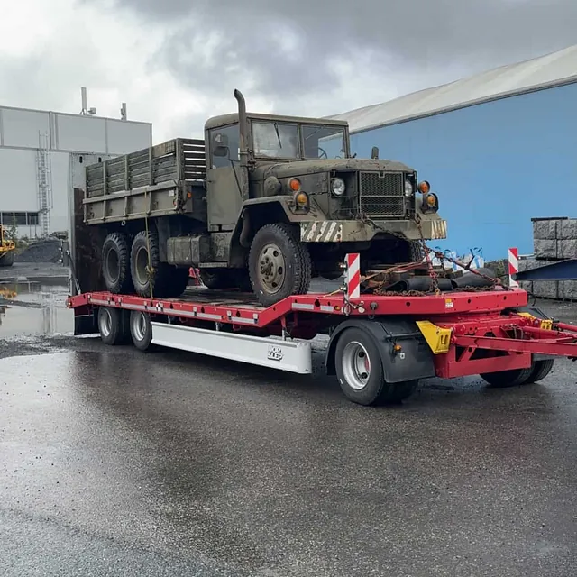 Militær lastebil på rød spesialhenger, parkert på våt asfalt utenfor industribygg.