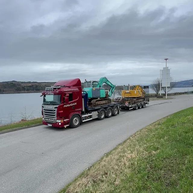 Rød Haugan-lastebil transporterer to beltegående gravemaskiner langs vei ved vann, under grå himmel.