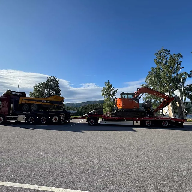Rød Haugan-lastebil med tilhenger transporterer dumper og oransje gravemaskin, parkert ved rasteplass omgitt av skog og innsjø.