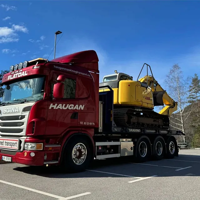 Rød Haugan-lastebil med bred transport-skilt frakter gul gravemaskin på parkeringsplass omgitt av fjell og trær.