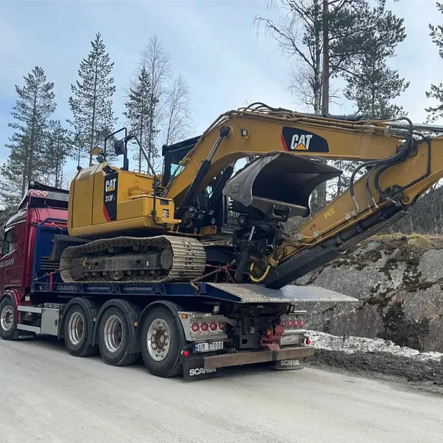 Rød Haugan-lastebil frakter en stor gul Caterpillar 318F beltegraver på spesialhenger langs vinterpreget vei.