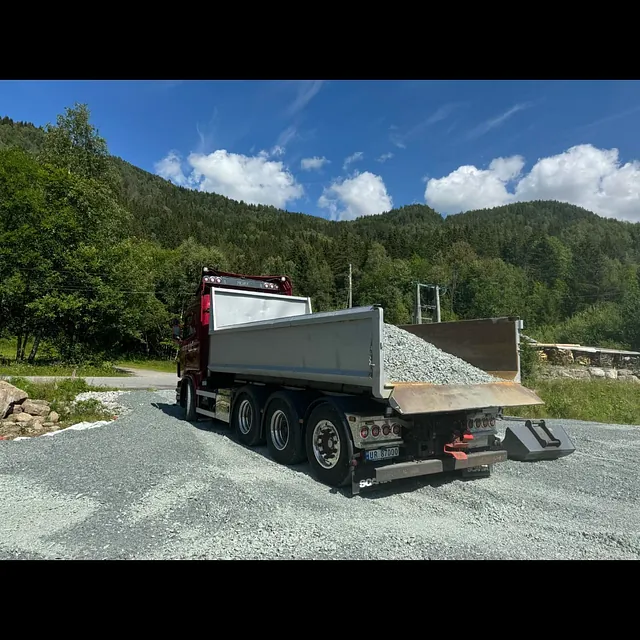 Tippbil fra Haugan med gruslast, avlasting på grusvei i skogsområde med blå himmel.