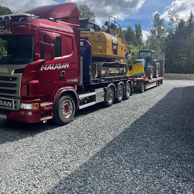 Rød Haugan-lastebil frakter gravemaskin og traktor med frontlaster på spesialhenger, parkert på grusplass under blå himmel.