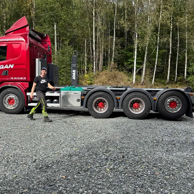 Rød trekkvogn fra Haugan med sjåfør foran, parkert på grusplass med skog i bakgrunnen.