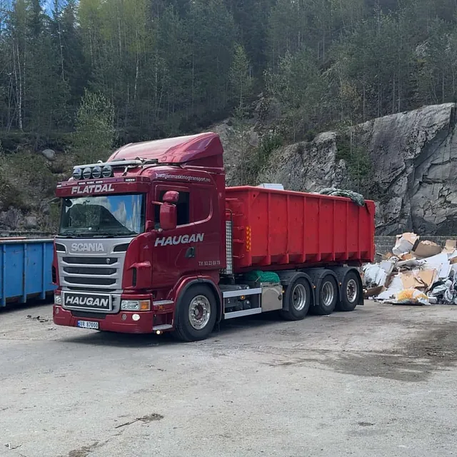 Rød Haugan-lastebil med rød container parkert ved gjenvinningsstasjon omgitt av papp og avfall.