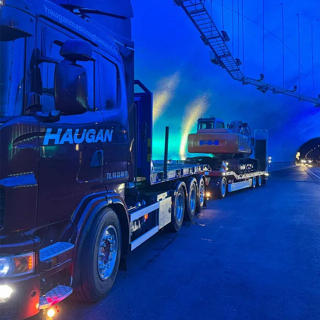 Lastebil med gravemaskin på henger kjører gjennom opplyst tunnel med blått lys.